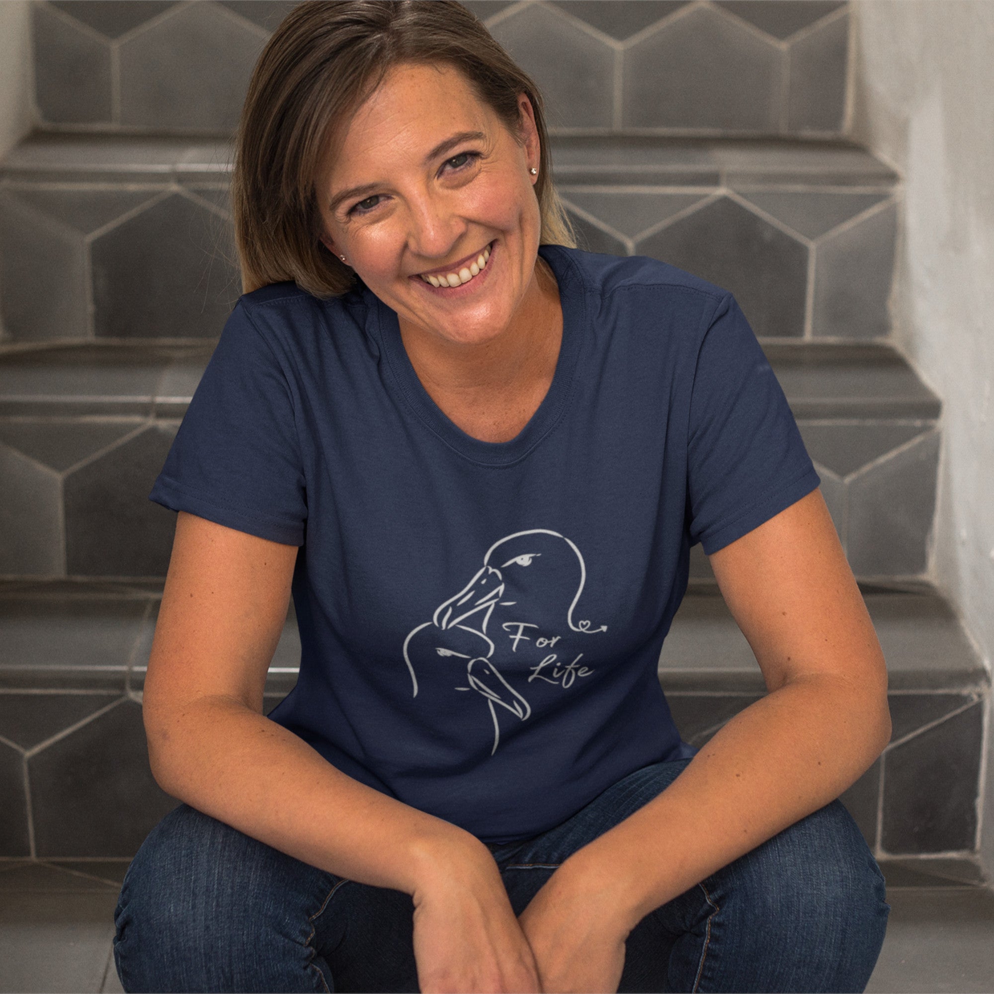 Navy blue unisex bird shirt featuring a line drawing of two Albatross heads and the words "For Life" on the front. Worn by a happy woman sitting on some stairs.