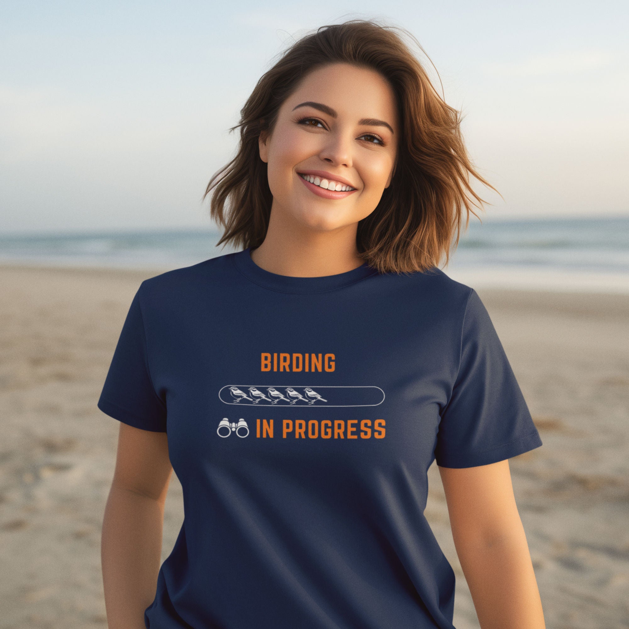 Navy blue bird shirt with the words Birding in Progress on the front around a loading progress bar, worn by a woman on the beach.