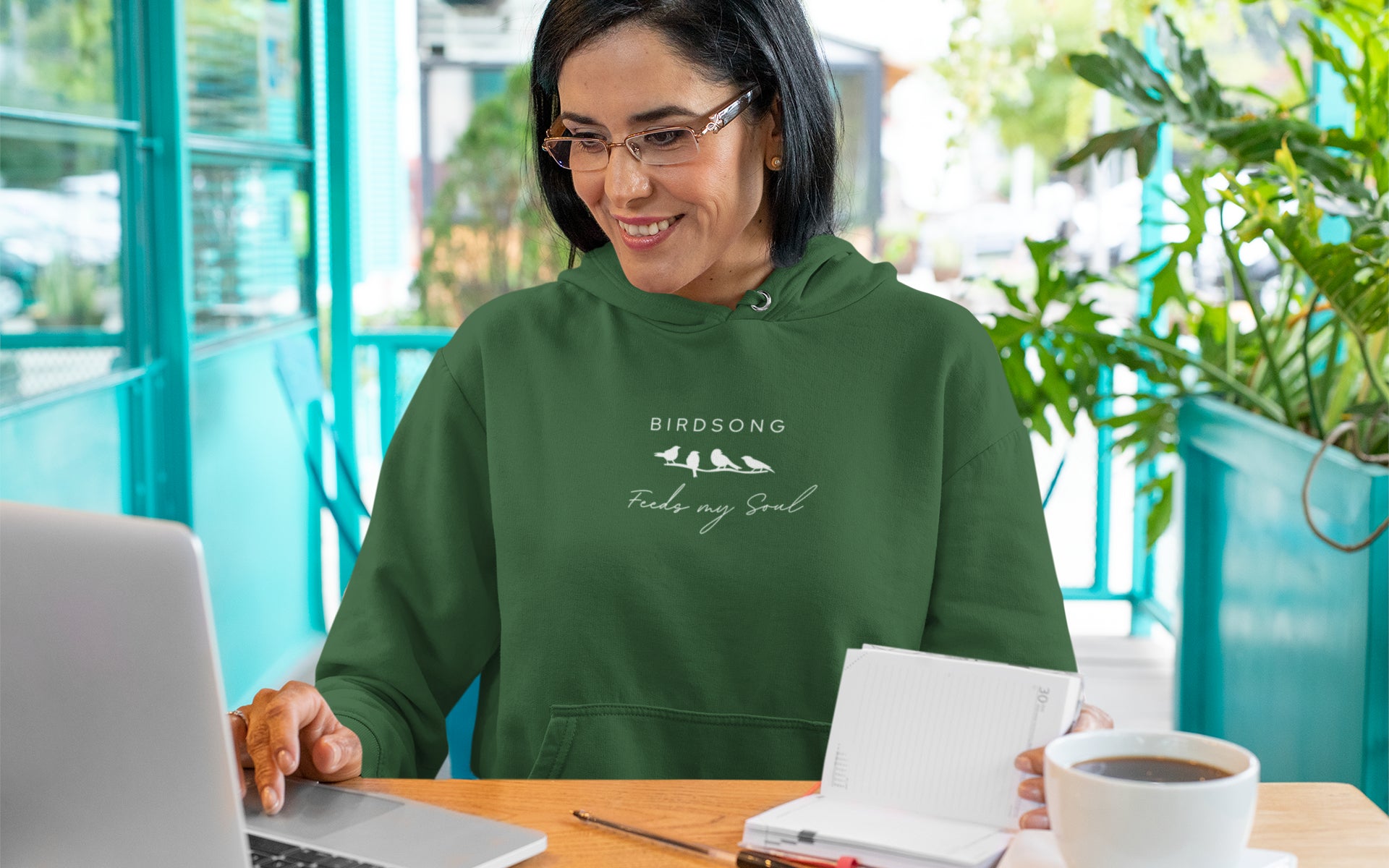 Forest green hoodie with the words Birdsong feeds my should above and below a white graphic of 4 birds on a branch. Worn by a woman working at a computer.