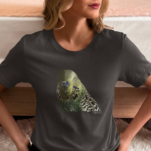 Dark gray unisex t-shirt with an illustrated and lifelike graphic of a green and yellow parakeet or budgie on the front. Below and to the right of the picture is the word Budgerigar and below that the scientific name in parenthesis in small print. Worn by a woman sitting on the floor.