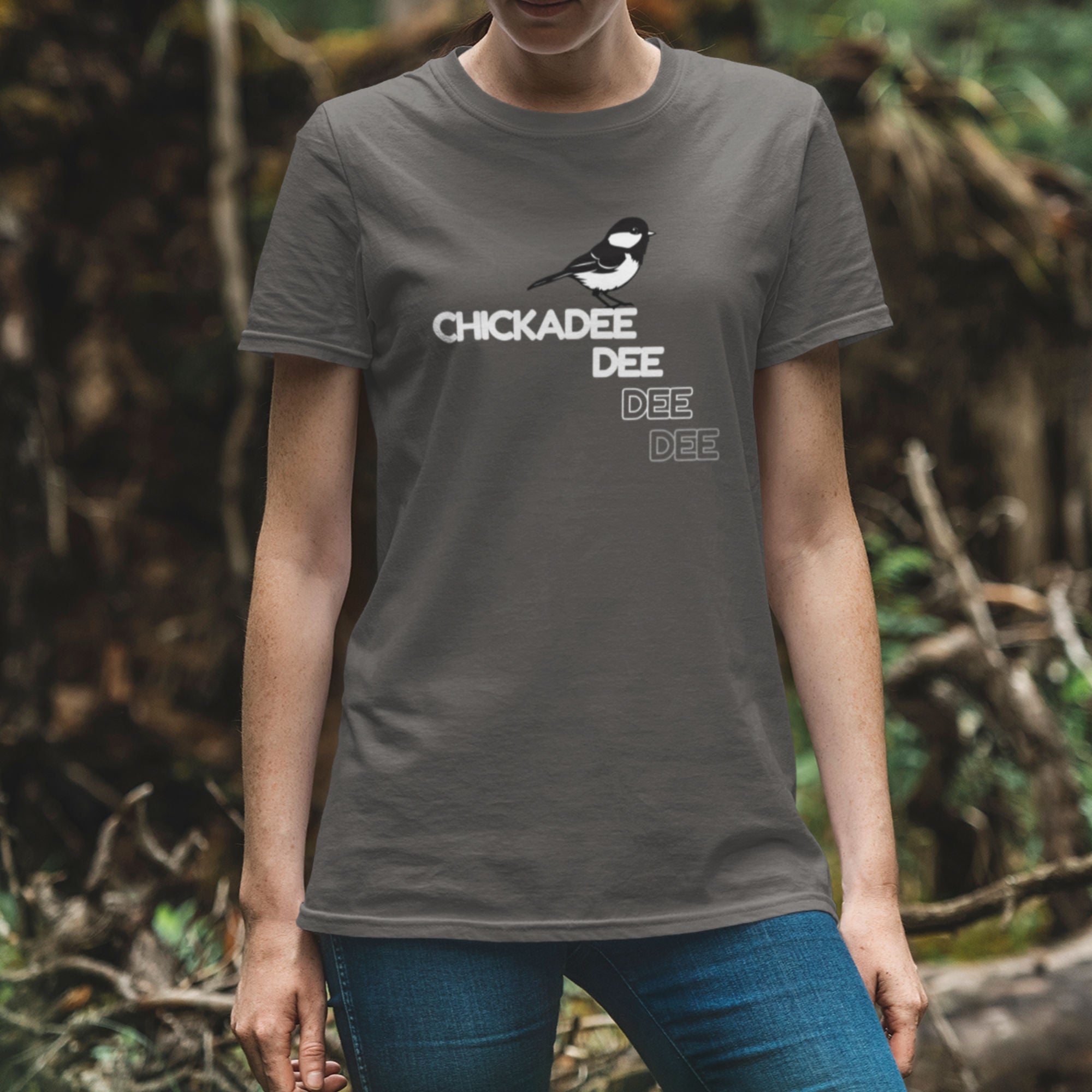 Grey birder shirt with a Chickadee and the words Chickadee dee dee dee on the front. Worn by a woman in the woods.