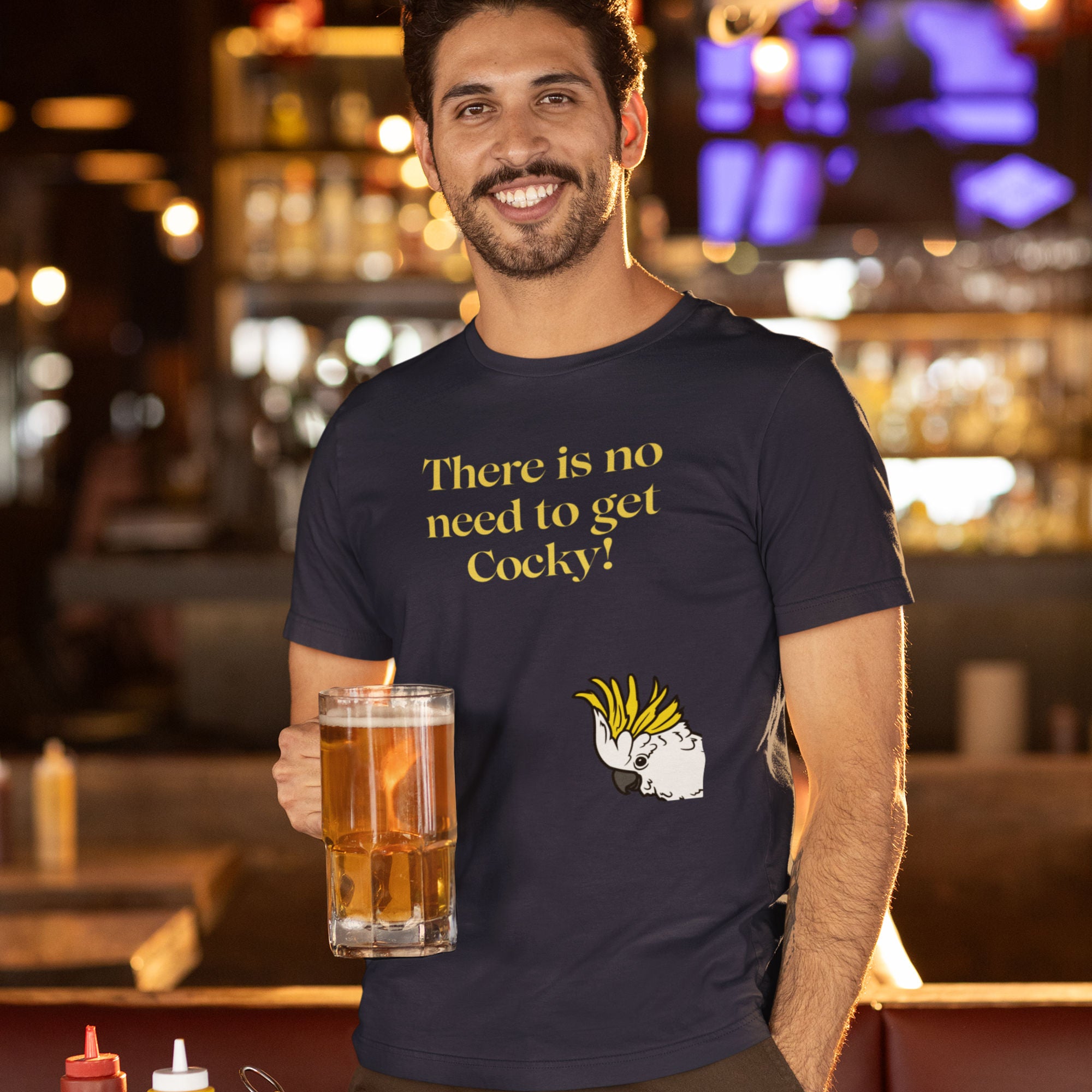 Navy blue bird shirt with the words, "There is no need to get Cocky!" on the front with a sulfur crested cockatoo peeking in from the side. Worn by a man with a beer at a bar.