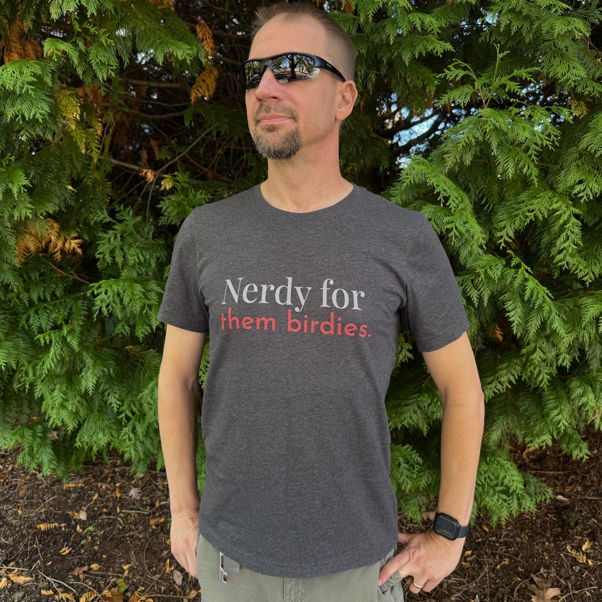 Dark grey unisex bird shirt with the words Nerdy for them birdies on the front in two coordinating fonts and colors. Worn by a man.