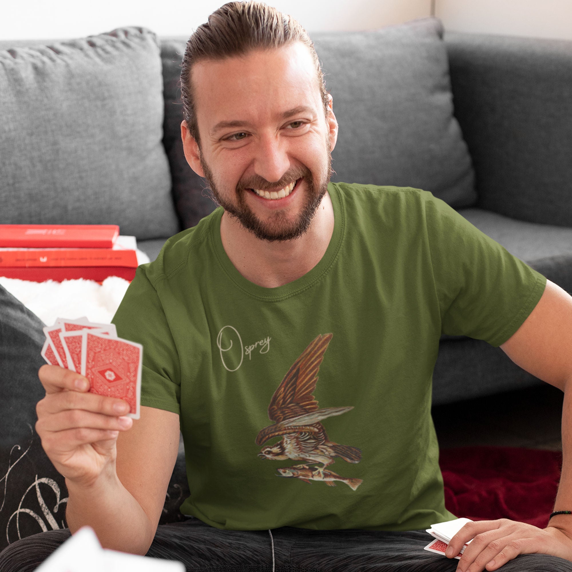 Olive green bird shirt with vintage art of an osprey with a fish in its talons and the word "Osprey in the top right. Worn by a man playing cards.