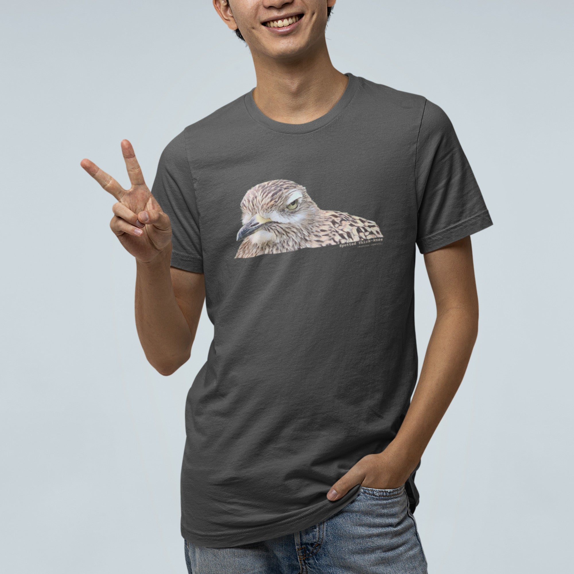 Dark gray unisex bird shirt with a lifelike illustration of a Spotted Thickknee on the front and the words Spotted Thickknee in small print below, with the scientific name included. Worn by a man giving the peace sign.