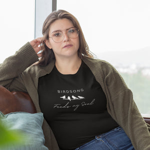 Black t-shirt with the words Birdsong feeds my soul placed above and below a white graphic of 4 birds on a branch. Worn by a woman relaxing on a couch.