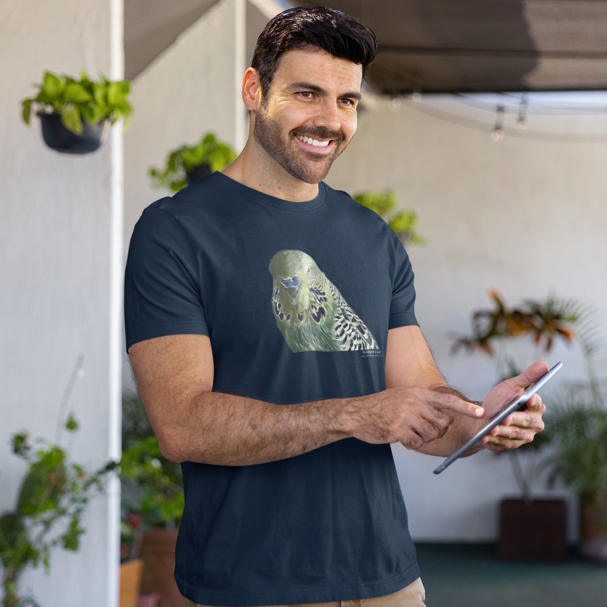 Navy unisex t-shirt with an illustrated and lifelike graphic of a green and yellow parakeet or budgie on the front. Below and to the right of the picture is the word Budgerigar and below that the scientific name in parenthesis in small print. Worn by a man holding an tablet.