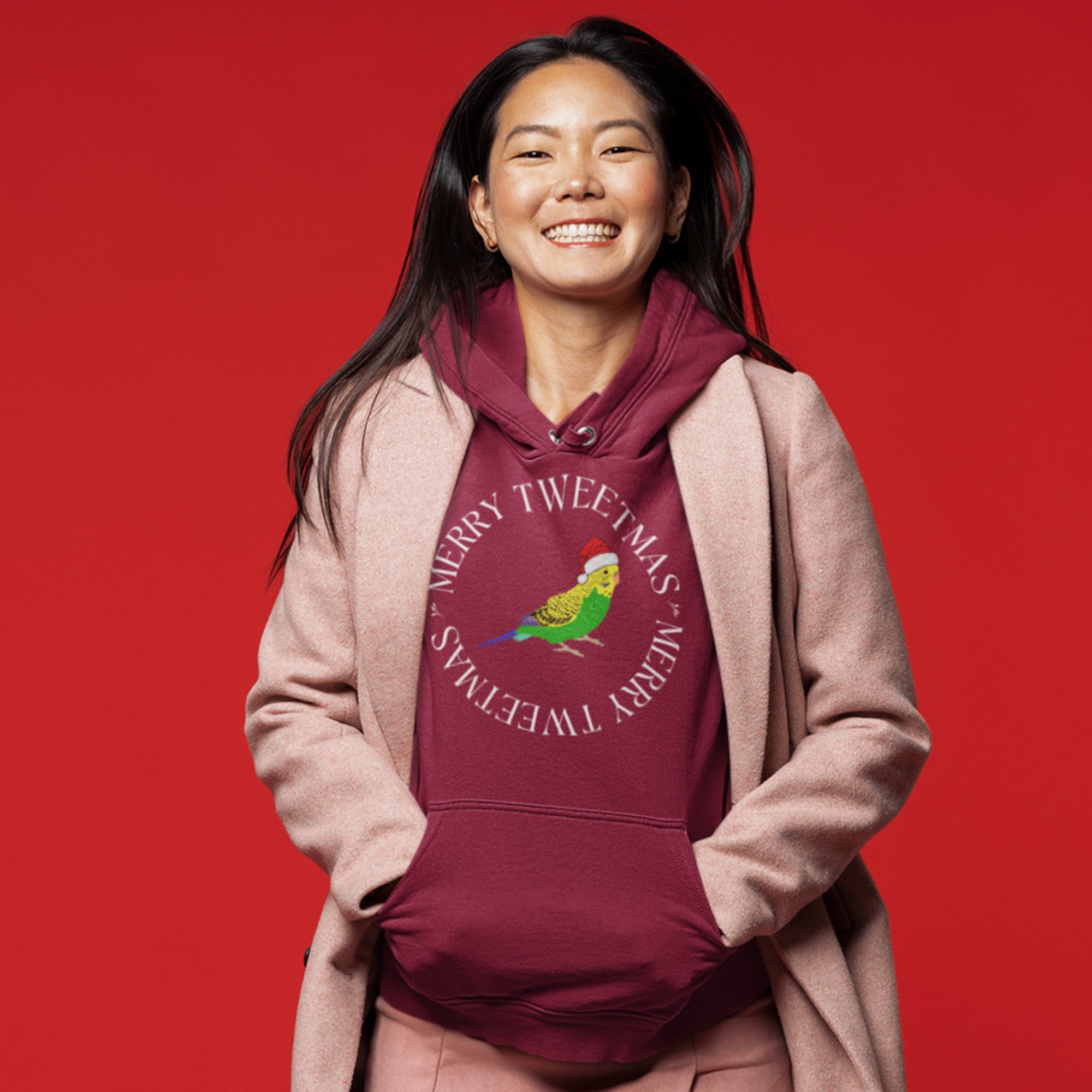 Maroon unisex hoodie with a budgie in a santa hat surrounded by the words Merry Tweetmas, separated by to seed sprigs. Worn by a woman.