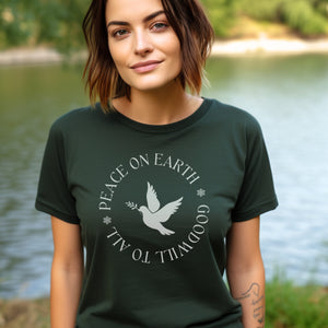 Forest green unisex bird t-shirt that says Peace on Earth Goodwill to All in a circle surrounding a dove of peace. Worn by a woman standing in front of a river.