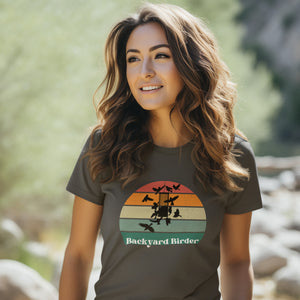 Dark gray bird shirt with a retro sun design outlining a bird feeder with the words Backyard Birder. Worn by a woman standing by a creek.