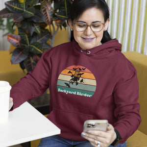 Maroon bird hoodie with a retro sun design outlining a bird feeder with the words Backyard Birder. Worn by a woman sitting in a waiting area.