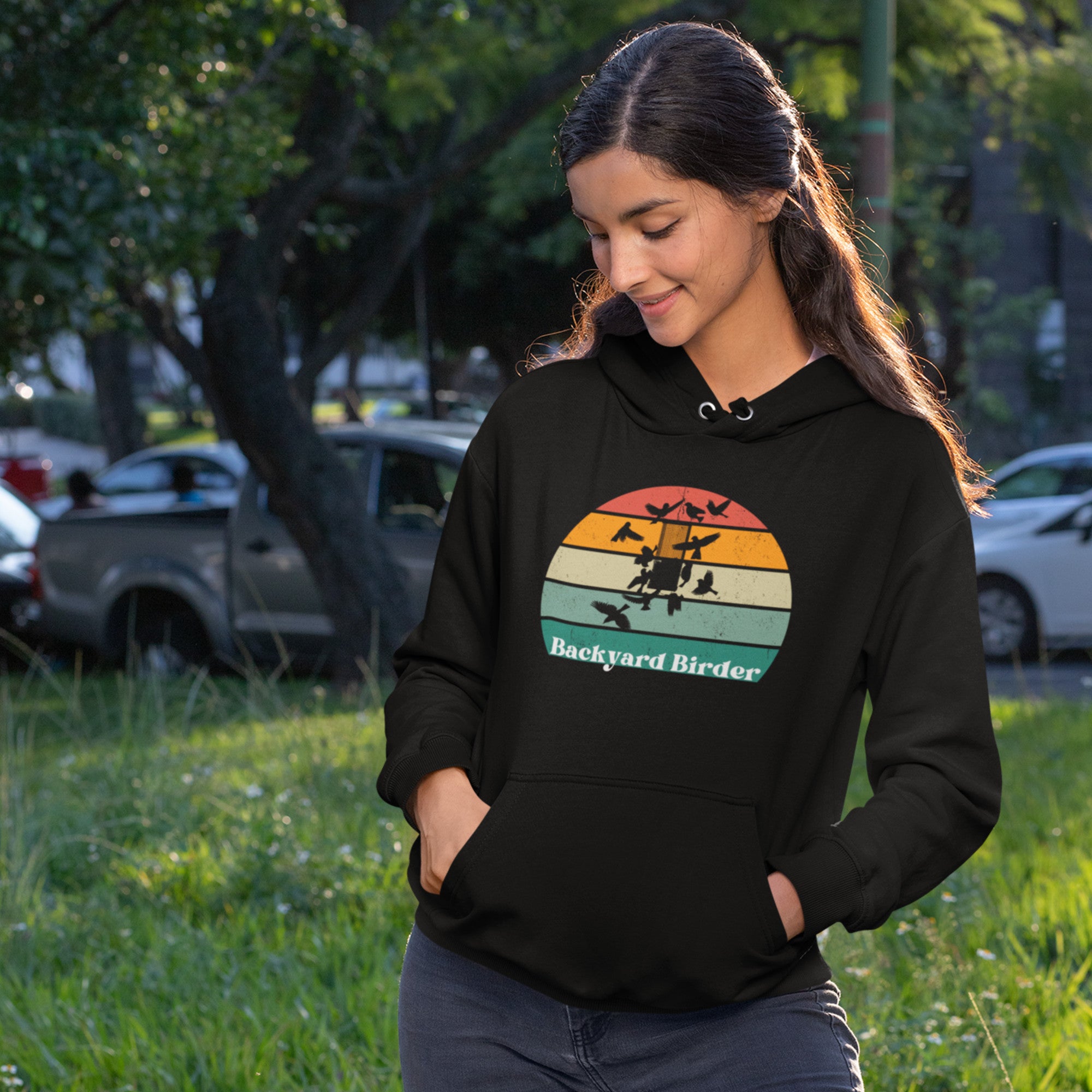 Black bird hoodie with a retro sun design outlining a bird feeder with the words Backyard Birder. Worn by a woman in the park.