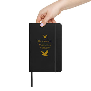 Hardcover bound birding notebook in black with the words, "Hawkward Moments, Birdwatching Journal" on the front with silhouettes of birds above and below. With a hand for scale.