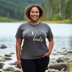 Grey unisex birding t-shirt with the words Love birds and a graphic of a bird on a branch in white. Worn by a woman standing by a river.