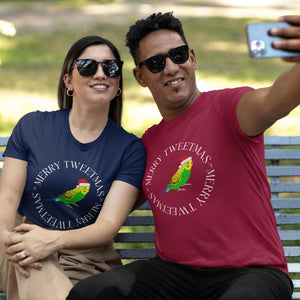 Navy blue and red unisex bird t-shirts with a budgie in a santa hat surrounded by the words Merry Tweetmas, separated by to seed sprigs. Worn by a couple sitting on a park bench taking a selfie. The woman is wearing navy blue and the man is wearing red.