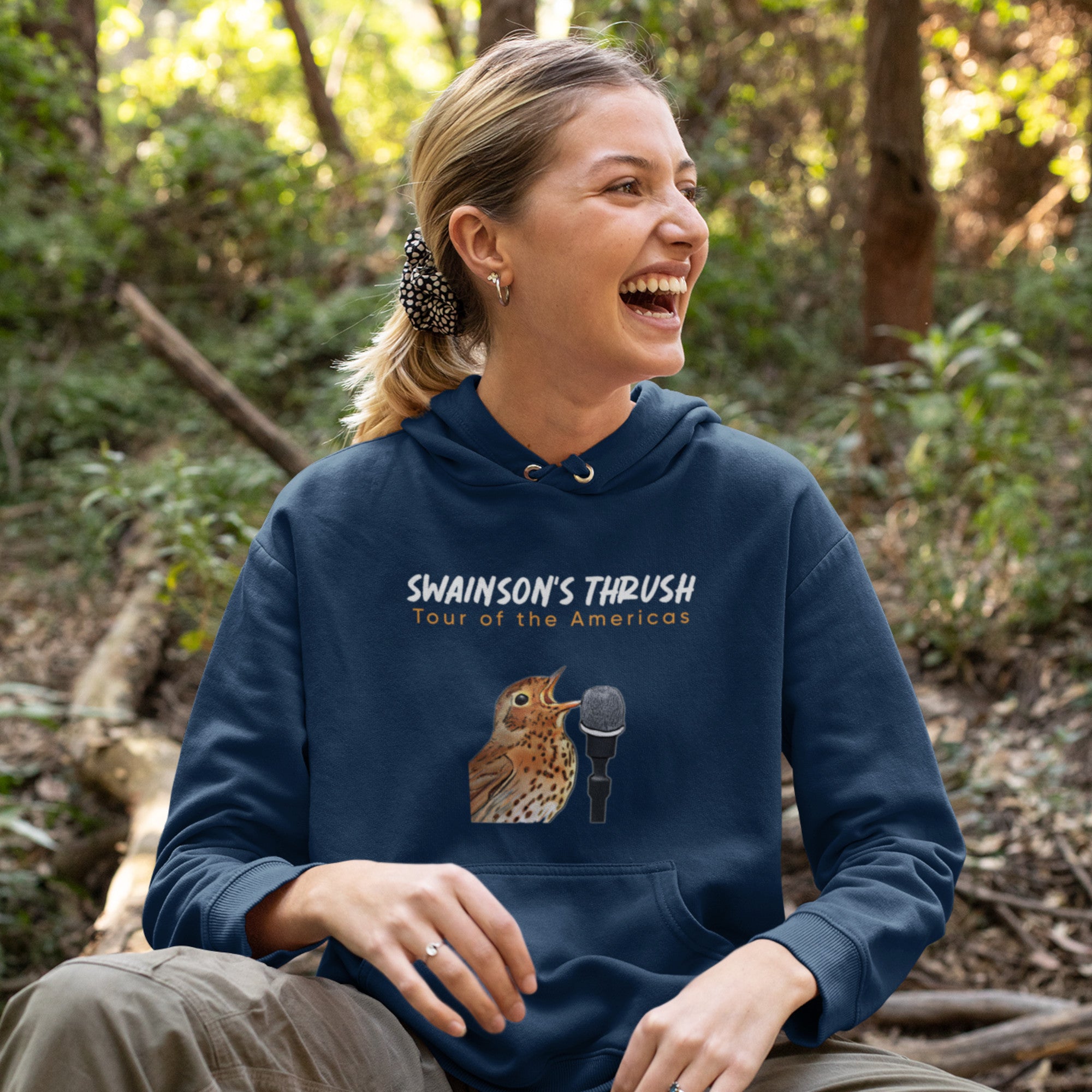 Navy blue unisex bird hoodie mimicking a concert hoodie with the words "Swainson's Thrush" and below that, "Tour of the Americas" and below that an illustrated Swainson's thrush singing into a microphone. Back of hoodie, (not shown) has "concert dates" and cities where this bird can be heard. Worn by a happy woman on a hike.