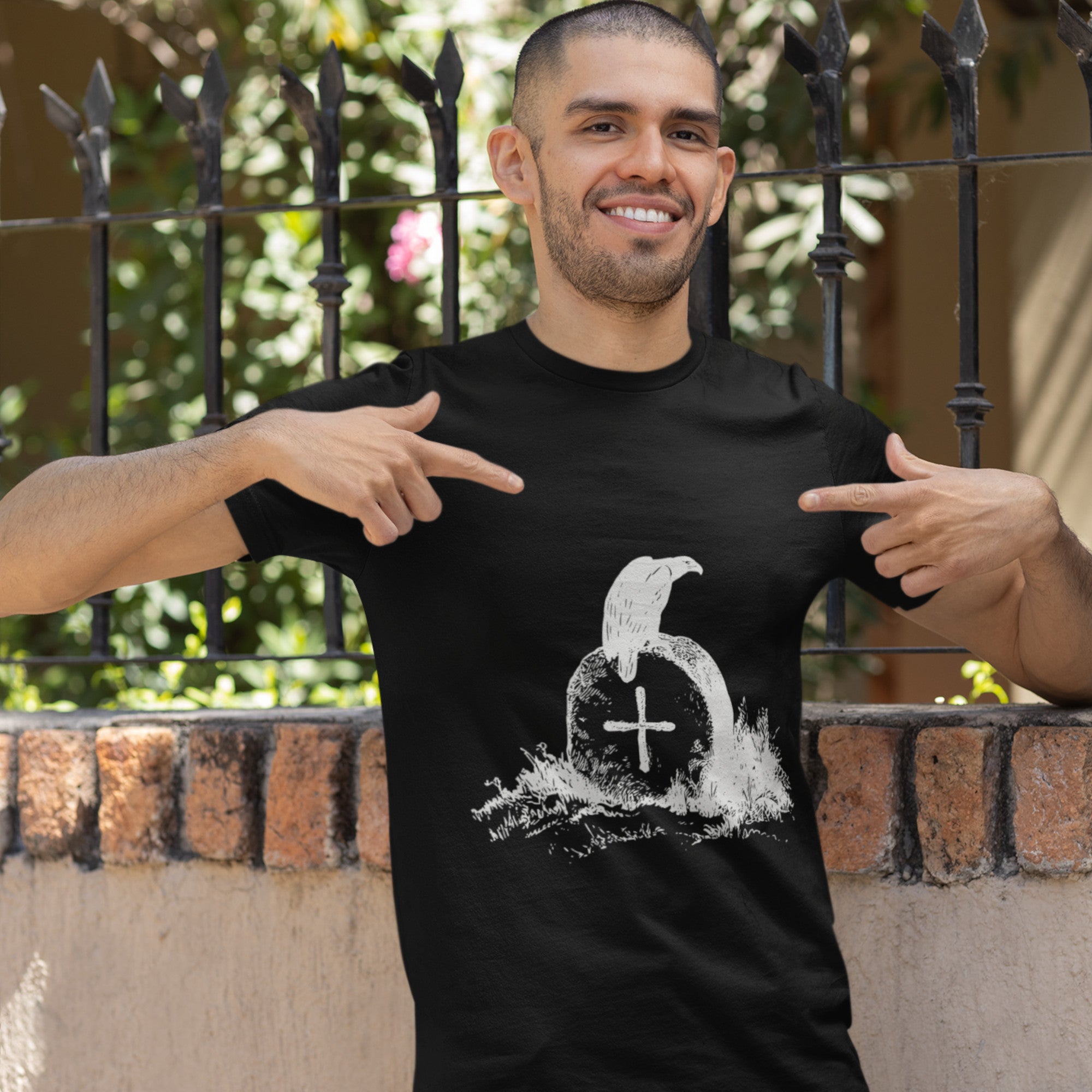 Black bird t-shirt with a picture of raven sitting on top of an unmarked grave. Worn by a man, looking happy and pointing to the front of the shirt.