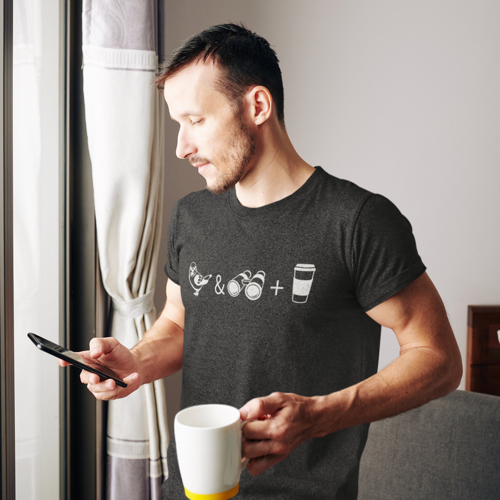 Man drinking coffee and looking at his phone while wearing a dark heather gray bird shirt with a bird, a pair of binoculars, and a to go coffee cup on the front.