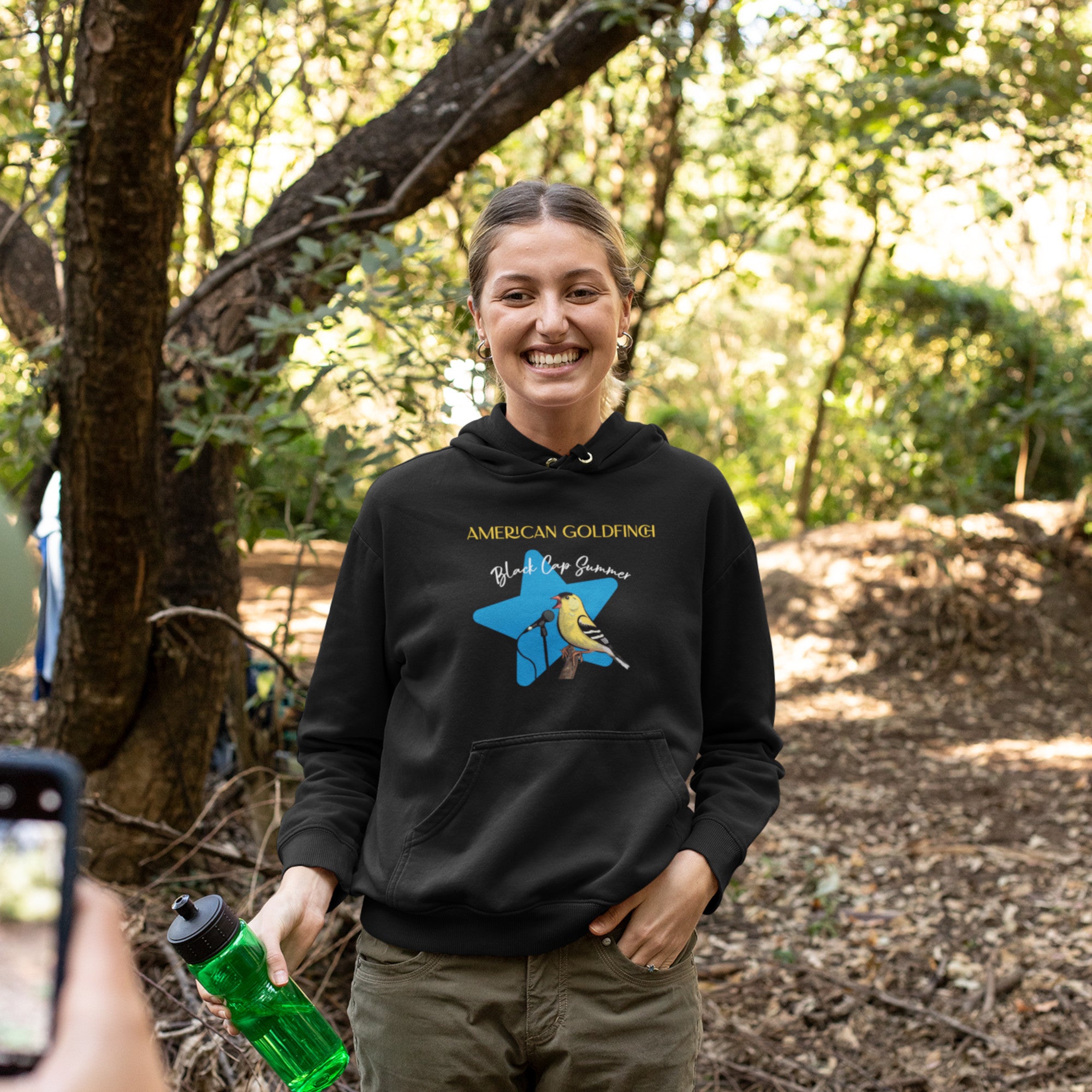 Black unisex bird hoodie featuring an American goldfinch singing into a microphone with a list of dates and cities on the back where the american goldfinch is commonly seen.