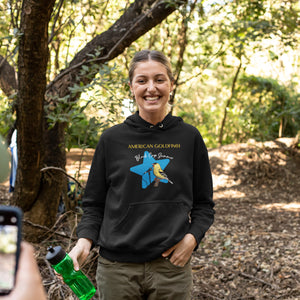 Black unisex bird hoodie featuring an American goldfinch singing into a microphone with a list of dates and cities on the back where the american goldfinch is commonly seen. Worn by a happy woman on a hike.