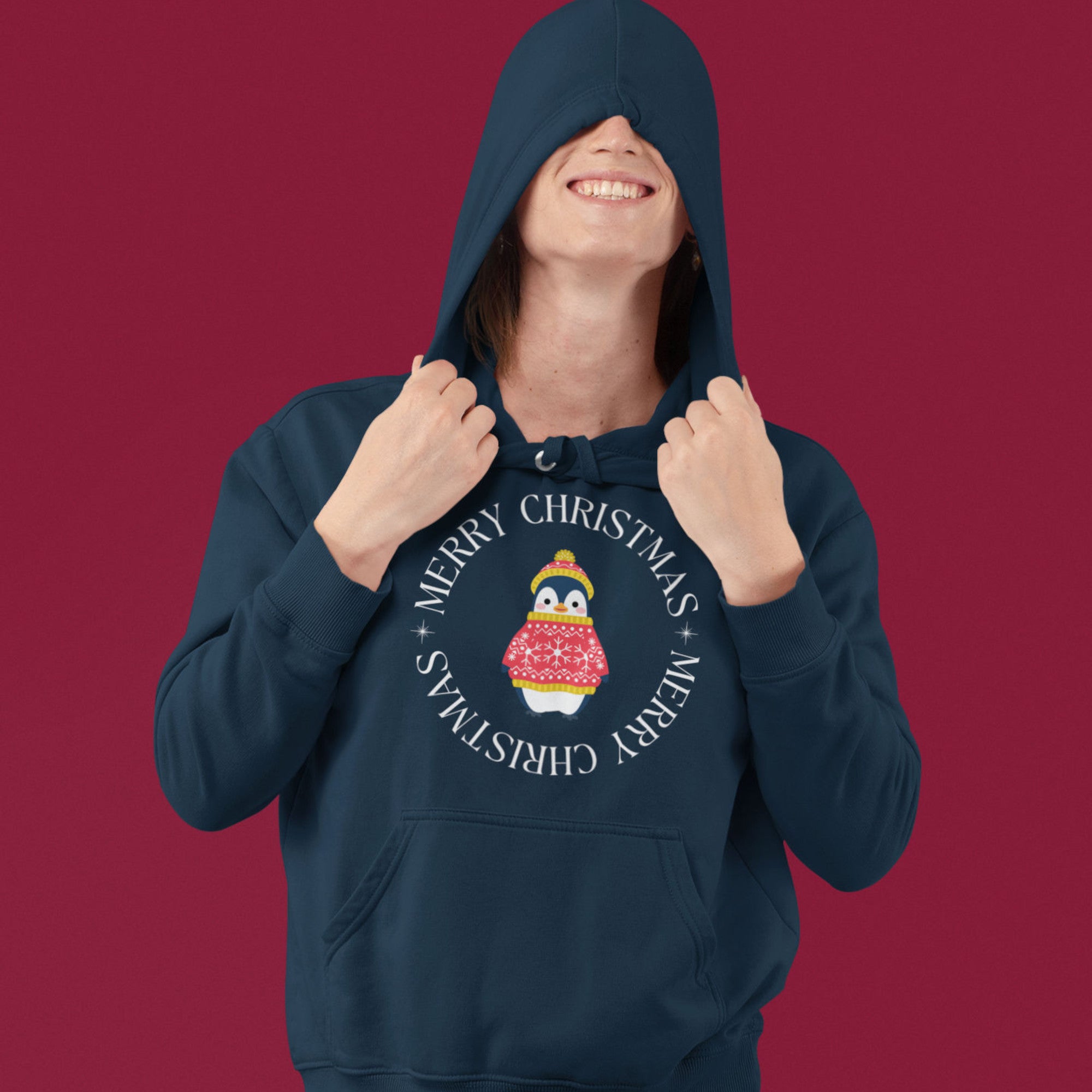 Navy blue unisex bird hoodie with Merry Christmas on the front in a circle that surrounds a cute penguin in a hat and a sweater. Worn by a person with the hood up.