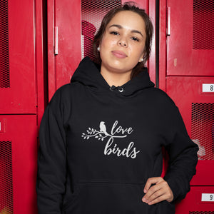 Black unisex bird hoodie with "love birds" on the front and a bird sitting on a branch. Worn by a woman standing in front of some lockers.