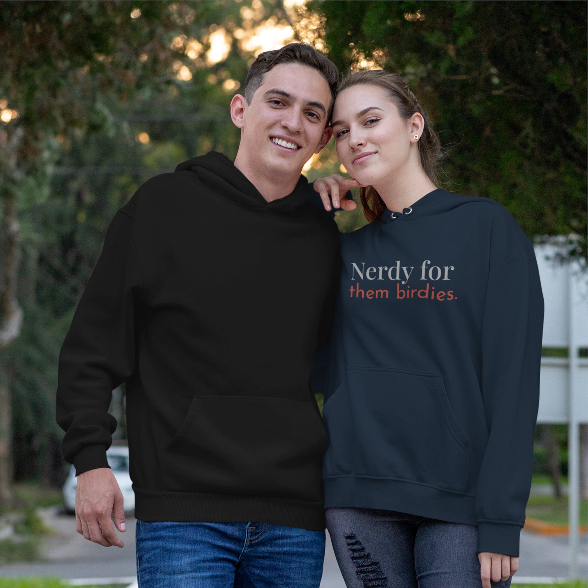 Navy blue unisex bird hoodie with the words Nerdy for them birdies on the front in two coordinating fonts and colors. Worn by a woman next to a man in a boring plain hoodie.