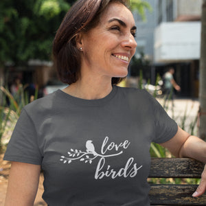 Grey unisex birding t-shirt with the words Love birds and a graphic of a bird on a branch in white. Worn by a woman sitting on a park bench.