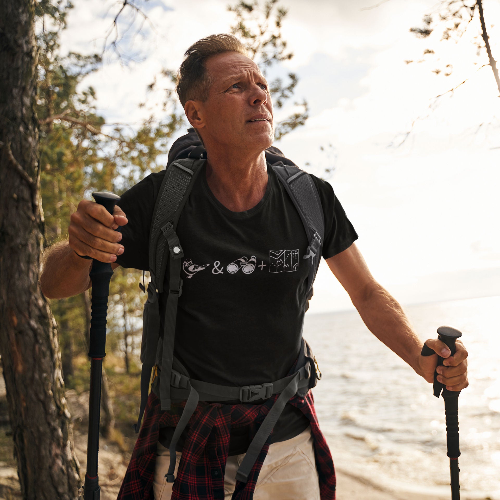 Unisex black bird t-shirt celebrating bird watching and the backcountry with a graphic of binoculars, a bird, and a map. Worn by a man on a hike.