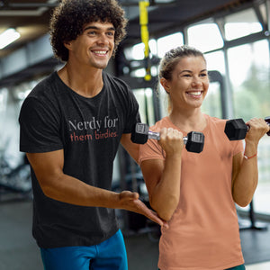 Black unisex bird shirt with the words Nerdy for them birdies on the front in two coordinating fonts and colors. Worn by a man coaching a woman in weightlifting form at the gym.