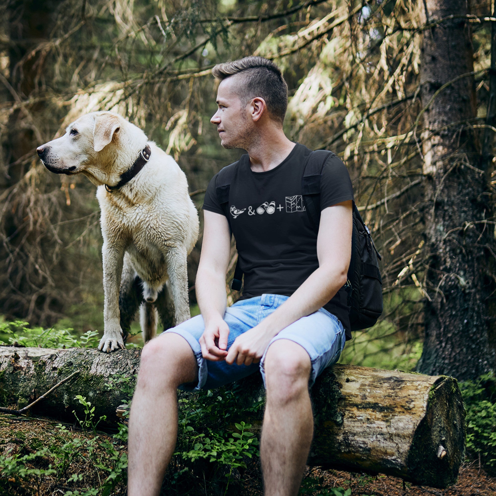 Unisex black bird t-shirt celebrating bird watching and the backcountry with a graphic of binoculars, a bird, and a map. Worn by a man on a hike with his dog, sitting on a log.