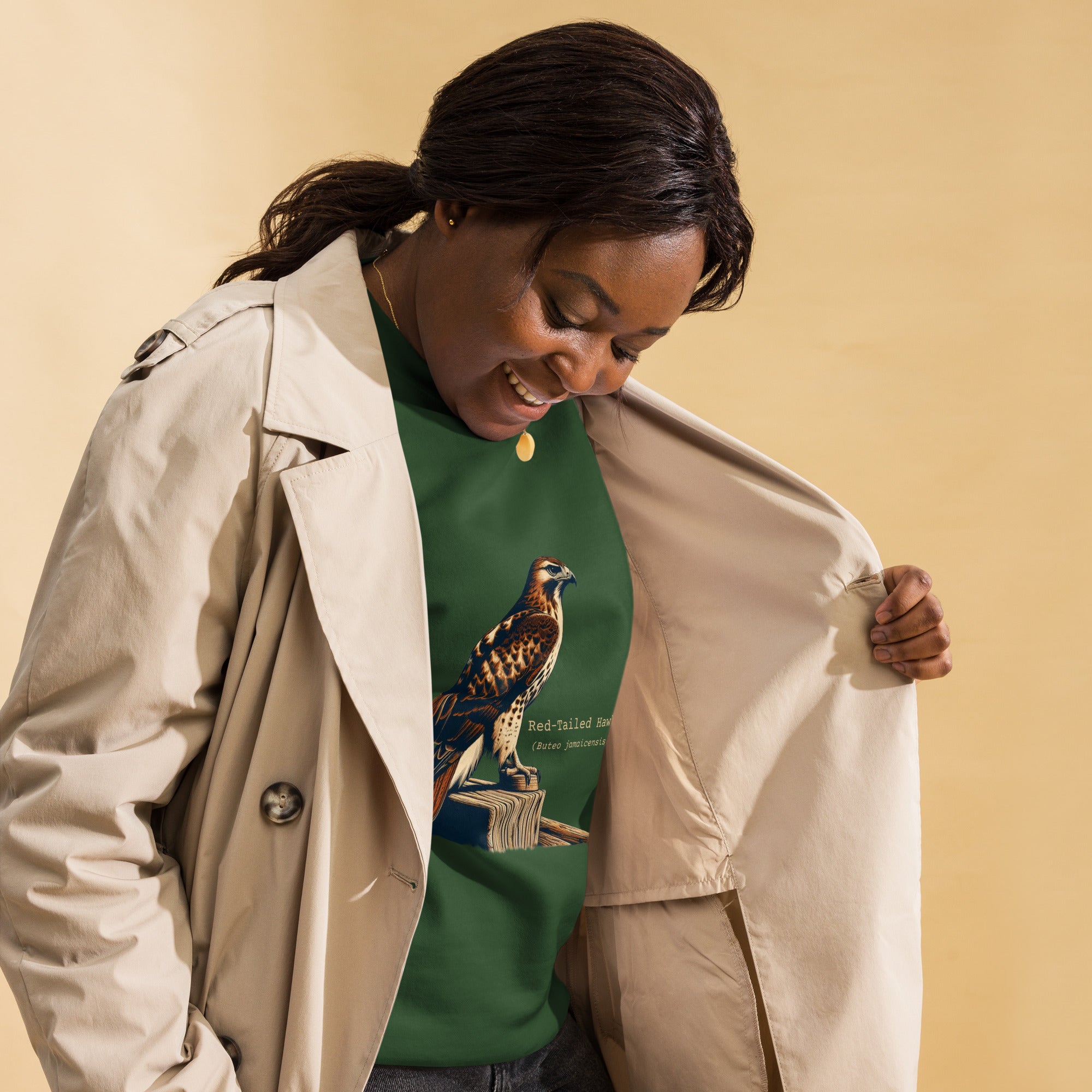 Green birding sweatshirt with an artistic depiction of a red-tailed hawk sitting on a fence and the common and scientific names for the bird next to it. Worn by a woman.