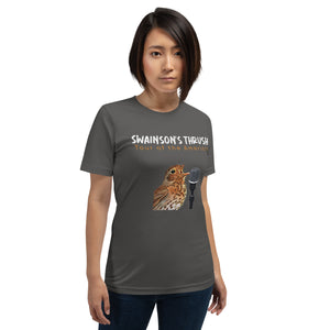 Dark gray unisex bird shirt mimicking a concert t-shirt with the words "Swainson's Thrush" and below that, "Tour of the Americas" and below that an illustrated Swainson's thrush singing into a microphone. Back of shirt, (not shown) has concert dates and cities where this bird can be heard. Worn by a woman.