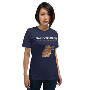 Navy unisex bird shirt mimicking a concert t-shirt with the words "Swainson's Thrush" and below that, "Tour of the Americas" and below that an illustrated Swainson's thrush singing into a microphone. Back of shirt, (not shown) has concert dates and cities where this bird can be heard. Worn by a woman.