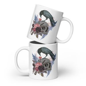 Gothic themed white 20-ounce bird mug with a crow carrying a heart pendant, perched on a skull surrounded by flowers, feathers and wings.