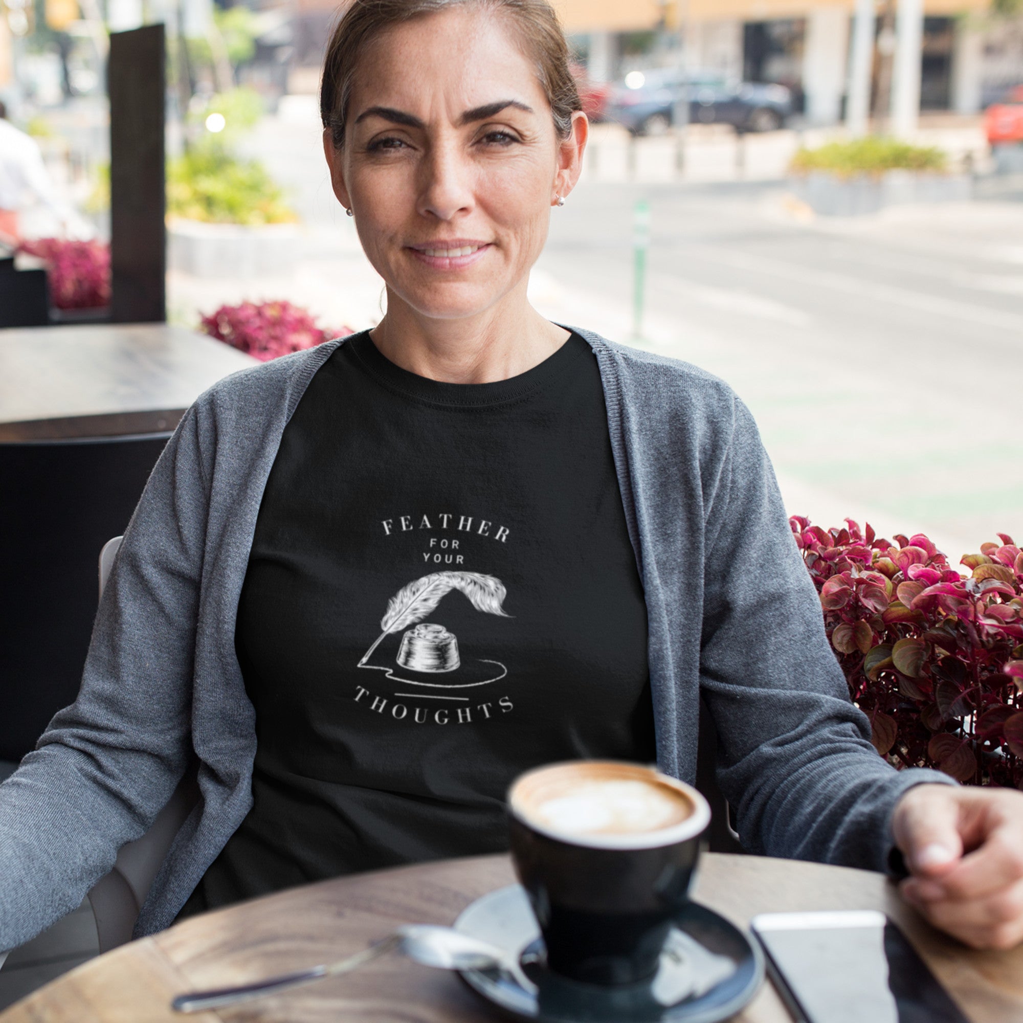 Black unisex t-shirt with a quill and an ink pot and the words, "Feather for your Thoughts." Worn by a woman having coffee at a cafe.