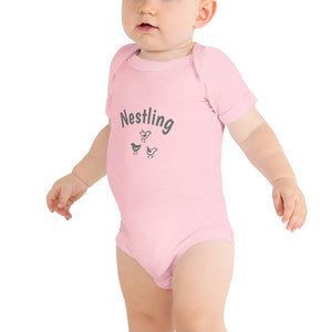 Pink one-piece baby suit with the word Nestling on the front above three small birds in dark grey. Worn by standing baby.