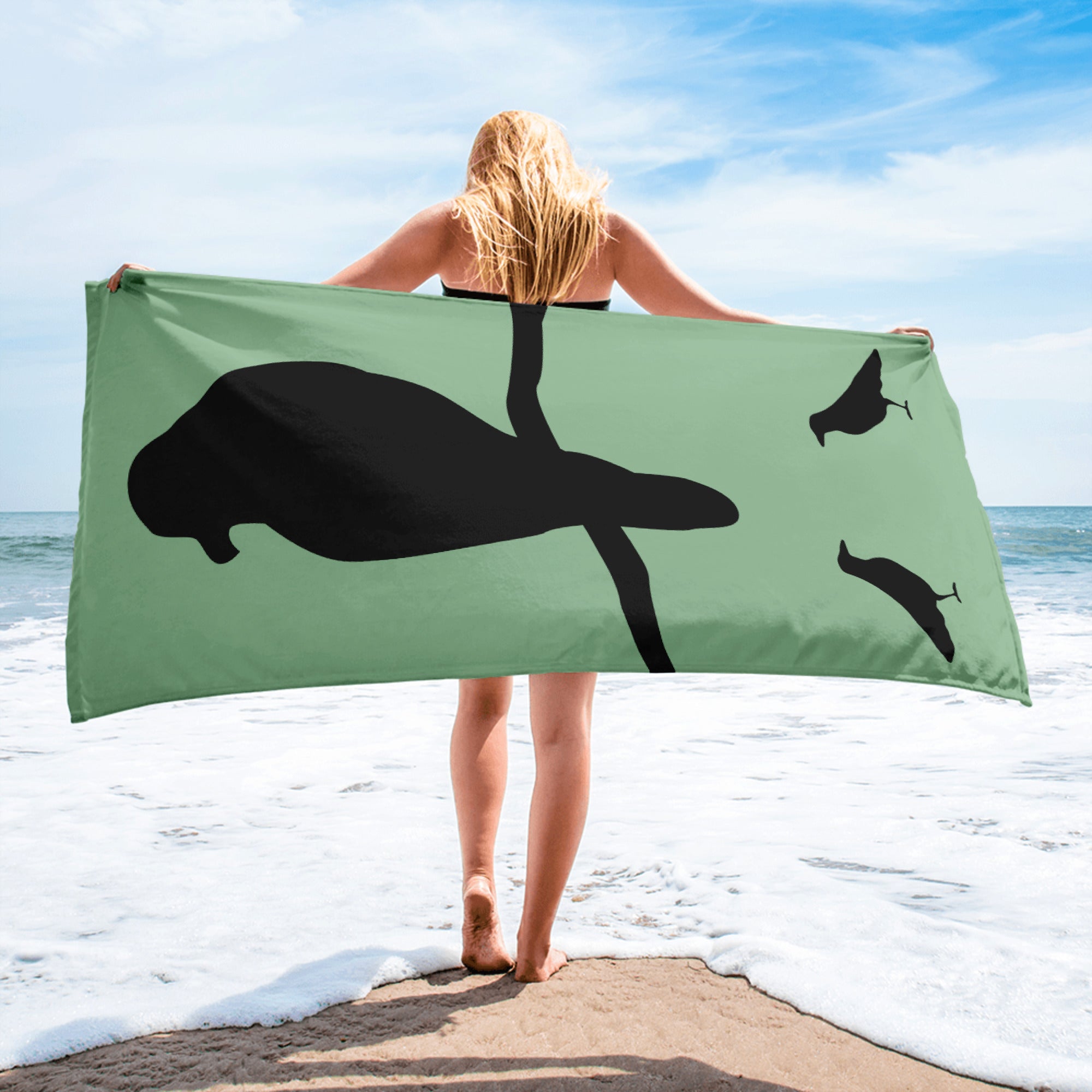 Sea green large beach towel with a large silhouette of a crow on a branch and two smaller standing crow silhouettes beneath, held by a blonde women while she walks on the beach.