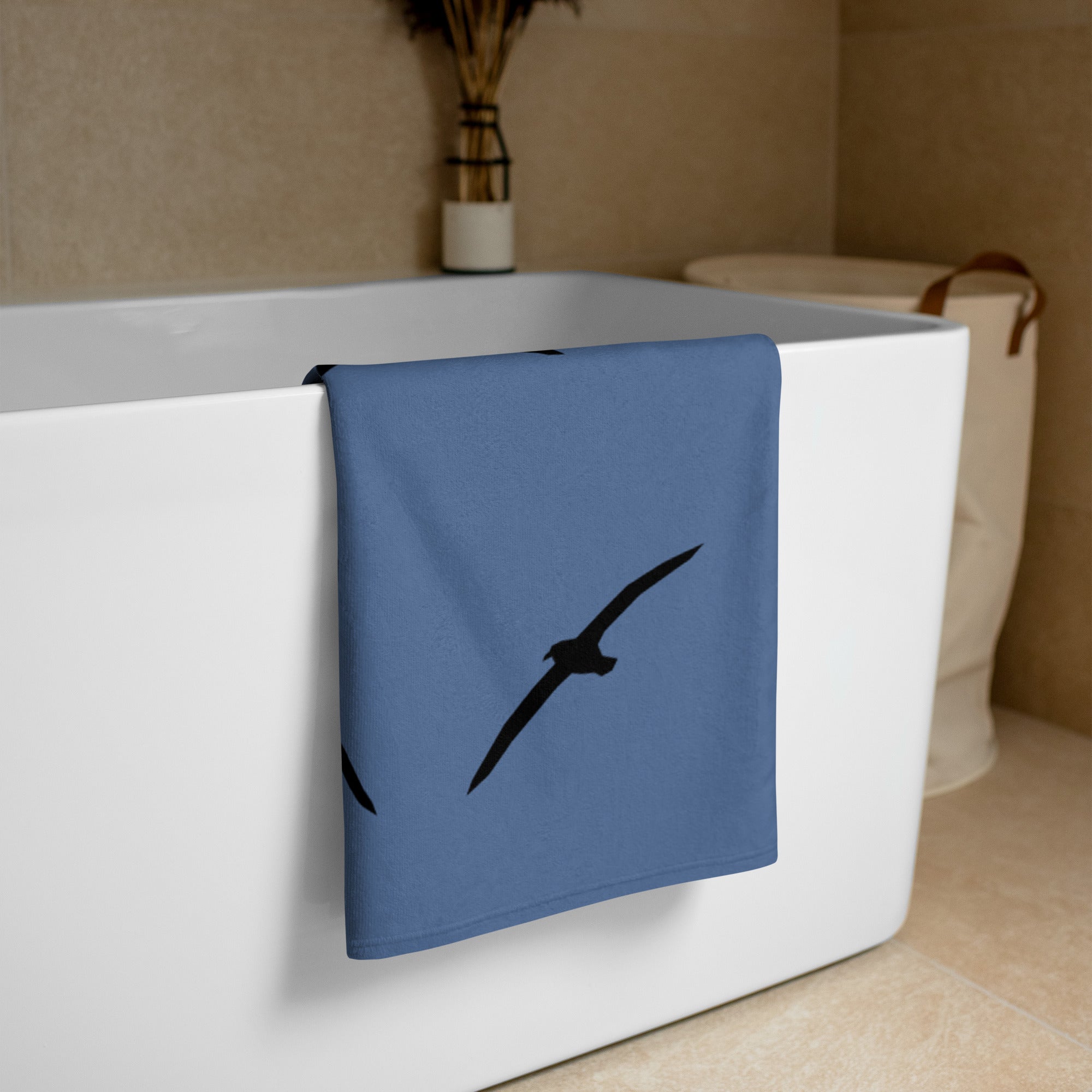 Blue large beach towel with a large silhouette of a sea gull on a branch and two smaller flying gull silhouettes beneath, folded and placed over the side of a free-standing bathtub.