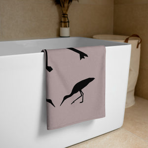 Dusty purple large beach towel with a large silhouette of a stork on a branch and two smaller stork silhouettes beneath, folded and placed over the side of a free-standing bathtub.