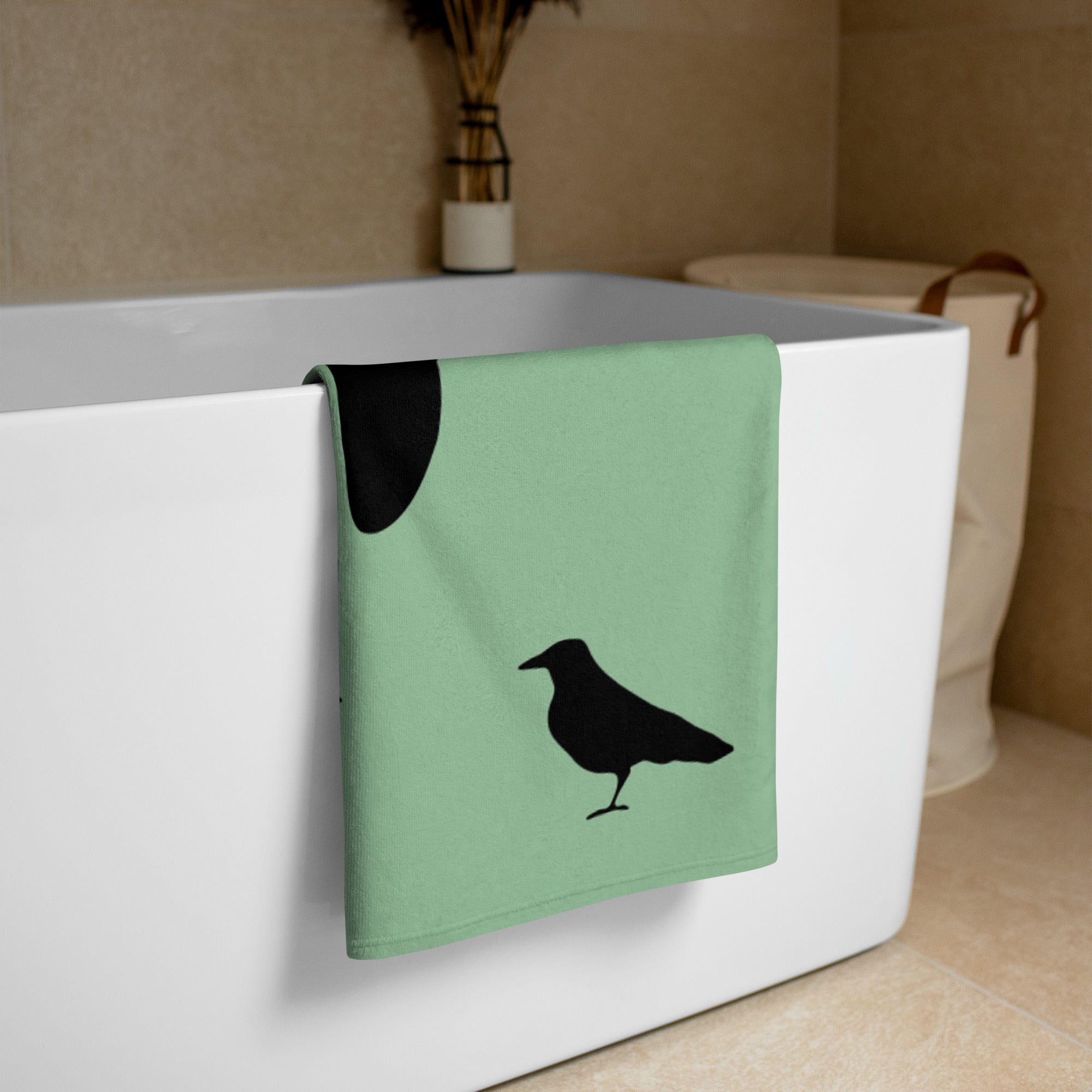 Sea green large beach towel with a large silhouette of a crow on a branch and two smaller standing crow silhouettes beneath, folded and placed over the side of a free-standing bathtub.