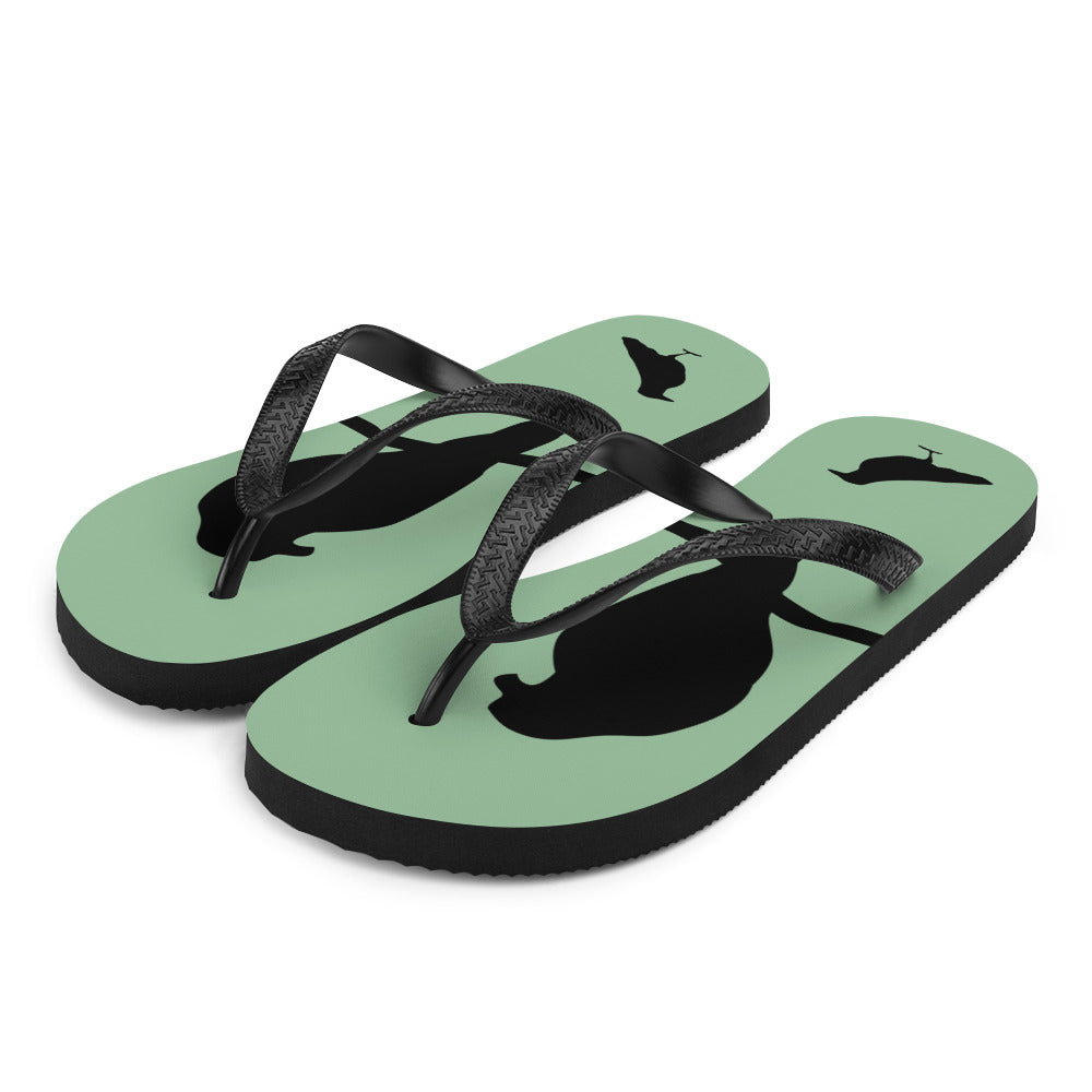 A pair of sea green flip-flops with black strap and a silhouette of a crow on a branch, viewed from the front at an angle..