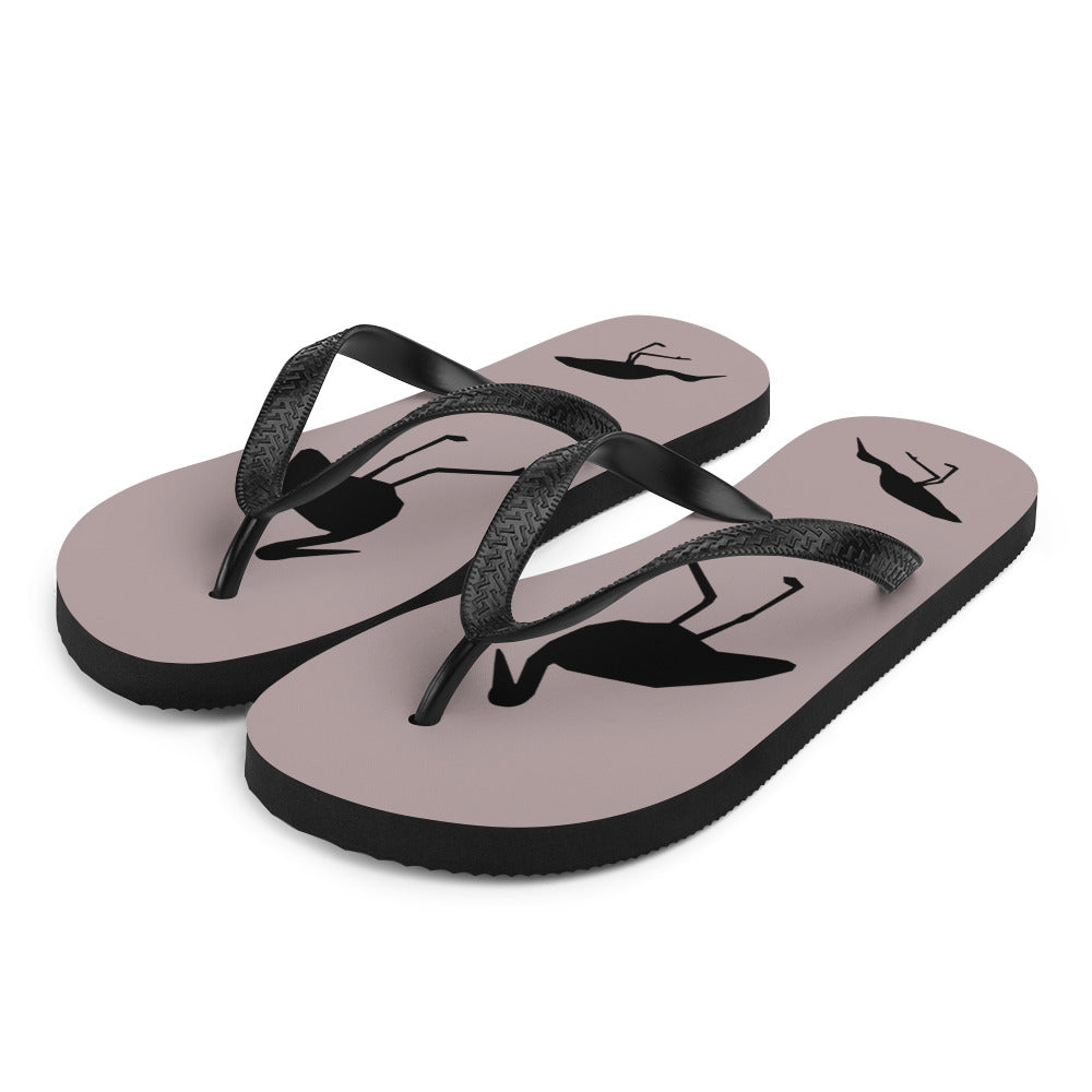 A pair of dusty purple flip-flops with black strap and a silhouette of a stork standing and smaller ones at the heel, viewed from the front left.