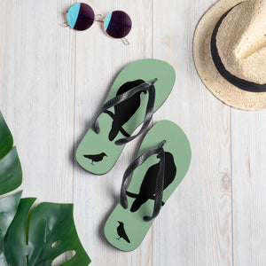 A pair of sea green flip-flops with black strap and a silhouette of a crow on a branch, with sunglasses and a hat.