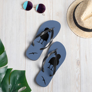 A pair of blue flip-flops with black strap and a silhouette of a seagull standing and smaller ones flying, with a hat and sunglasses.