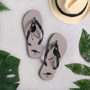 A pair of dusty purple flip-flops with black strap and a silhouette of a stork standing and smaller ones at the heel, viewed from the top down with a hat and some leaves nearby.