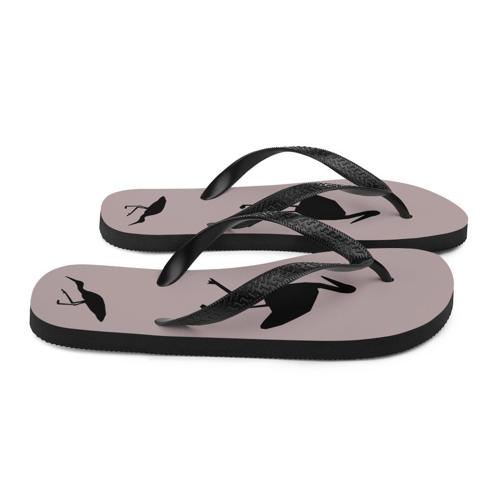 A pair of dusty purple flip-flops with black strap and a silhouette of a stork standing and smaller ones at the heel, viewed from the right side.