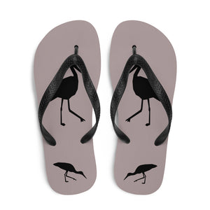 A pair of dusty purple flip-flops with black strap and a silhouette of a stork standing and smaller ones at the heel, viewed from the top down.