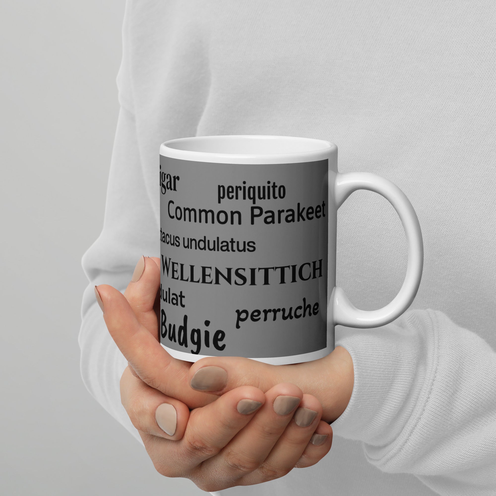 White 15 ounce coffee mug with an illustrated lifelike picture of a green and yellow budgie on a grey background next to the word budgerigar in other languages.  This view shows the words budgerigar, perruche, melopsittacus undulatus, and budgie.