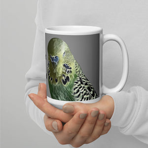 White 15 ounce coffee mug with an illustrated lifelike picture of a green and yellow budgie on a grey background and next to the word budgerigar in other languages.  This view shows the illustrated picture of the parakeet.